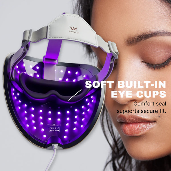 A close-up of a woman's face next to a wearable WAVLO Apollo SG LED light therapy mask. The mask is illuminated with purple light and has built-in eye cups, with text reading 'SOFT BUILT-IN EYE CUPS' and 'Comfort seal supports secure fit.