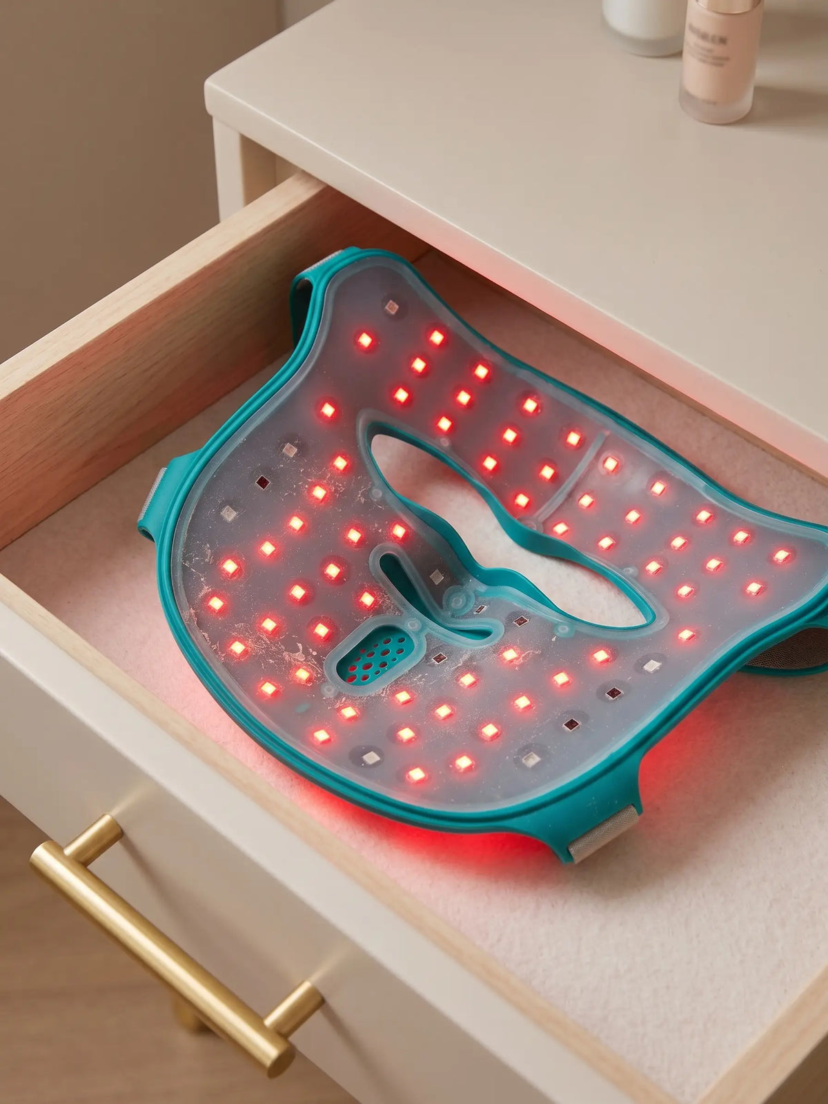 A teal LED light therapy face mask sits inside an open cream-colored drawer, with red lights illuminated across its surface.