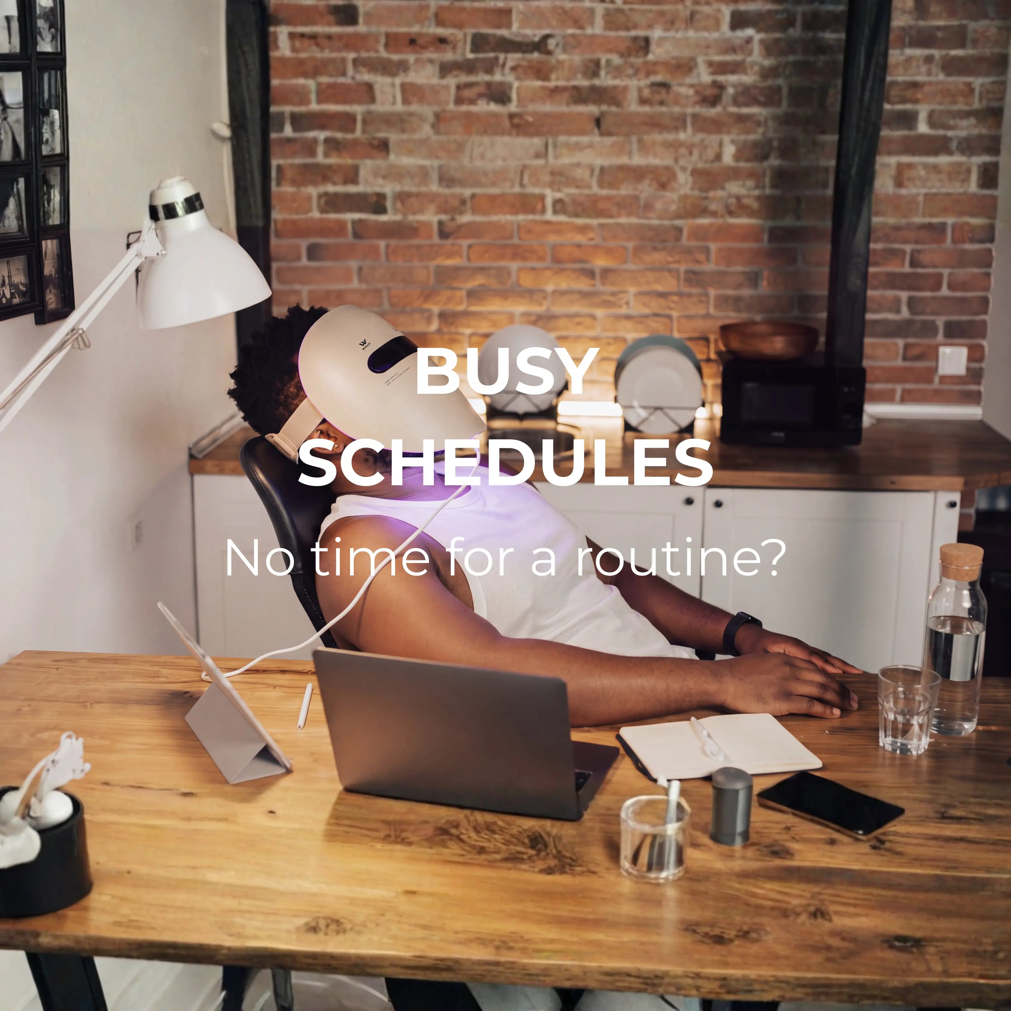 A person reclines on a chair at a wooden desk in a home office setting with a brick wall background, wearing a white led face mask - WAVLO Apollo SG for a blue and red light therapy for stress release. White text overlay reads 'BUSY SCHEDULES' and 'No time for a routine?'.