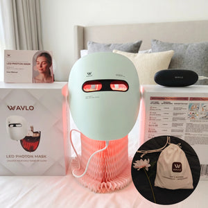 A light white LED face mask -WAVLO Apollo SG device with glowing red light panels, displayed on a bed alongside its white retail box, a black charging case, a storage bag, and an inset showing a branded drawstring pouch.