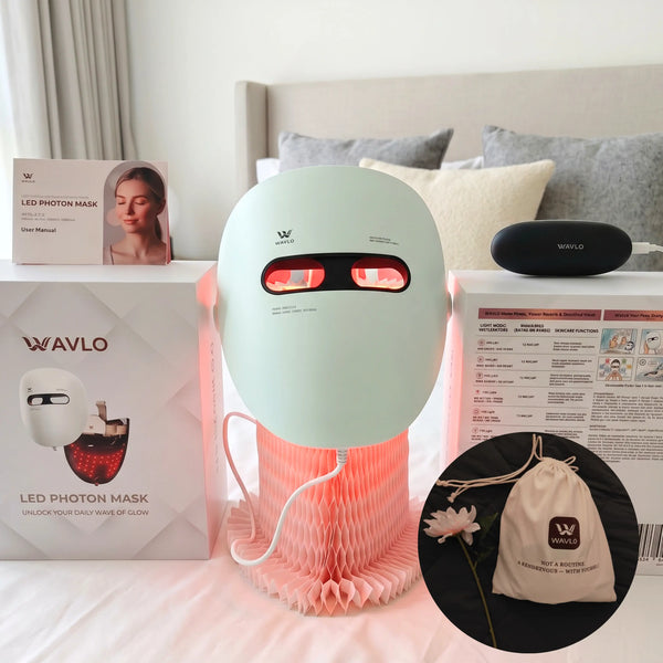 A light white LED face mask -WAVLO Apollo SG device with glowing red light panels, displayed on a bed alongside its white retail box, a black charging case, a storage bag, and an inset showing a branded drawstring pouch.