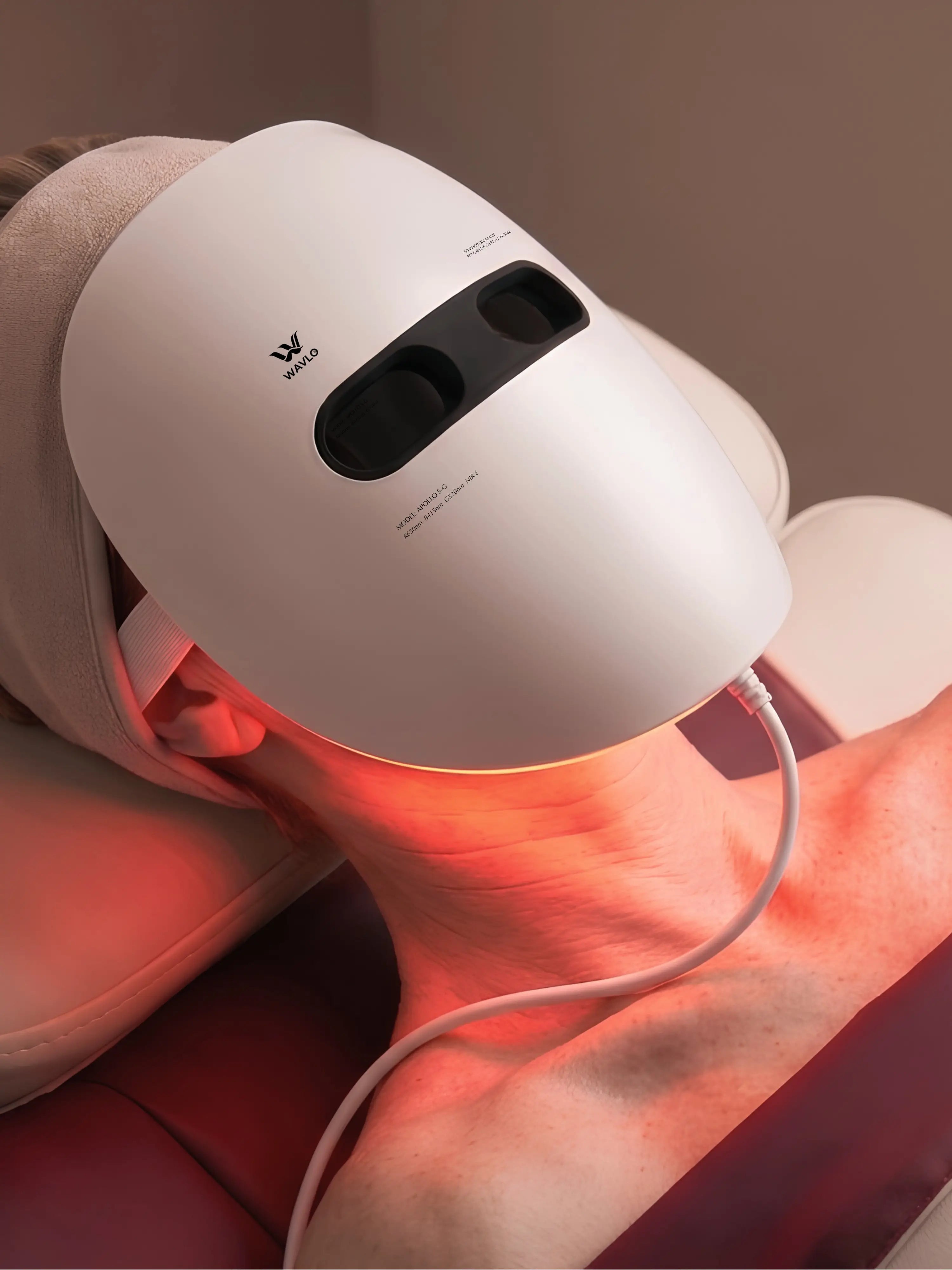 A white WAVLO Apollo SG LED neck and face treatment mask being worn by a person lying down, emitting a red light onto the skin.